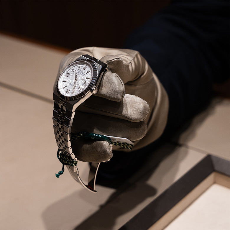 A gloved hand carefully holding a Rolex Land‑Dweller, angled forward in a presenting gesture as though being shown to a customer